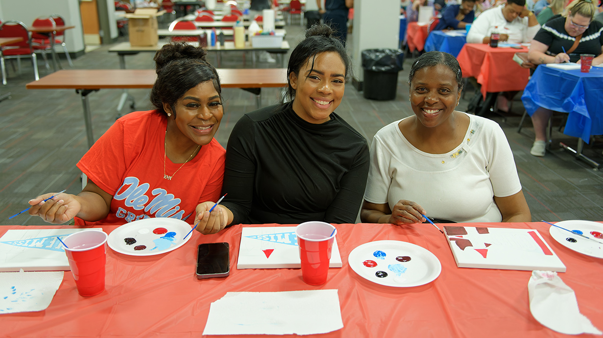 Three women smile as they pain on canvasses at a table topped with a red tablecloth.