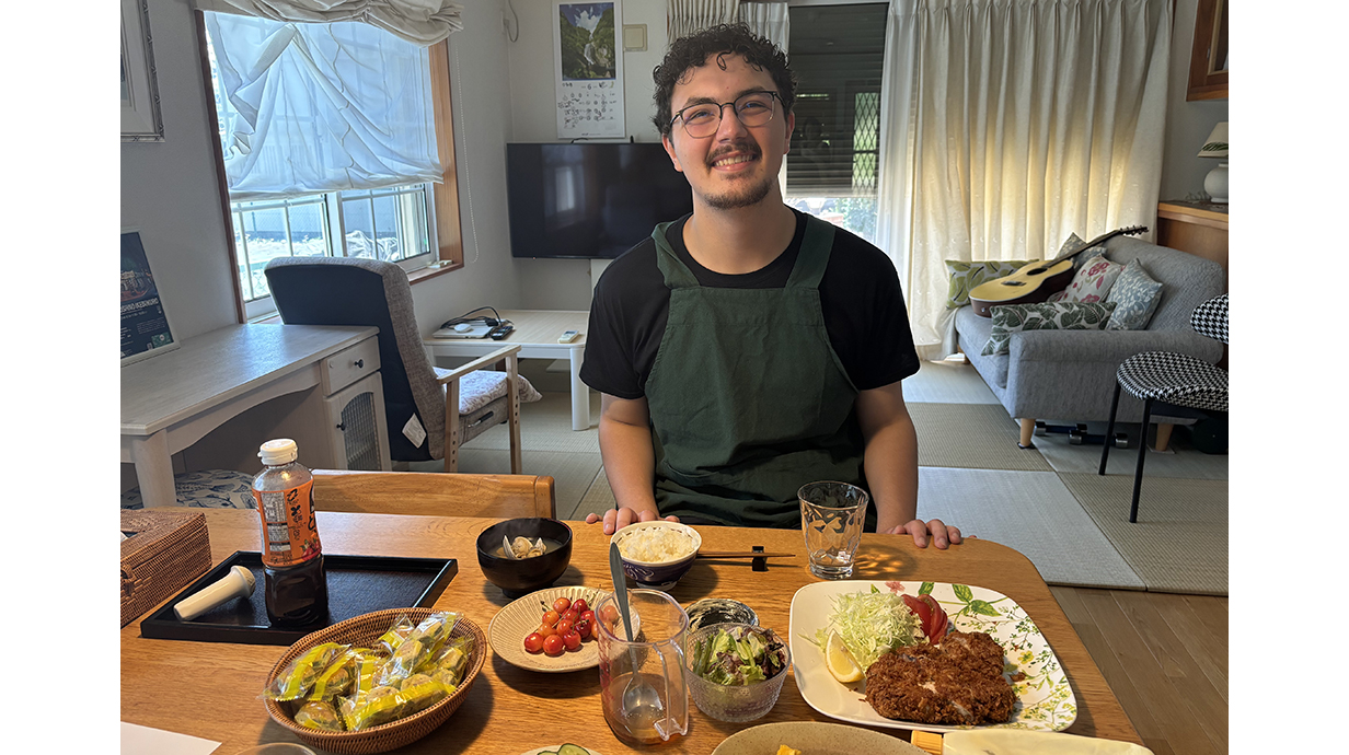 A young man wearing an apron sits at a table lined with bowls and plates of different foods.