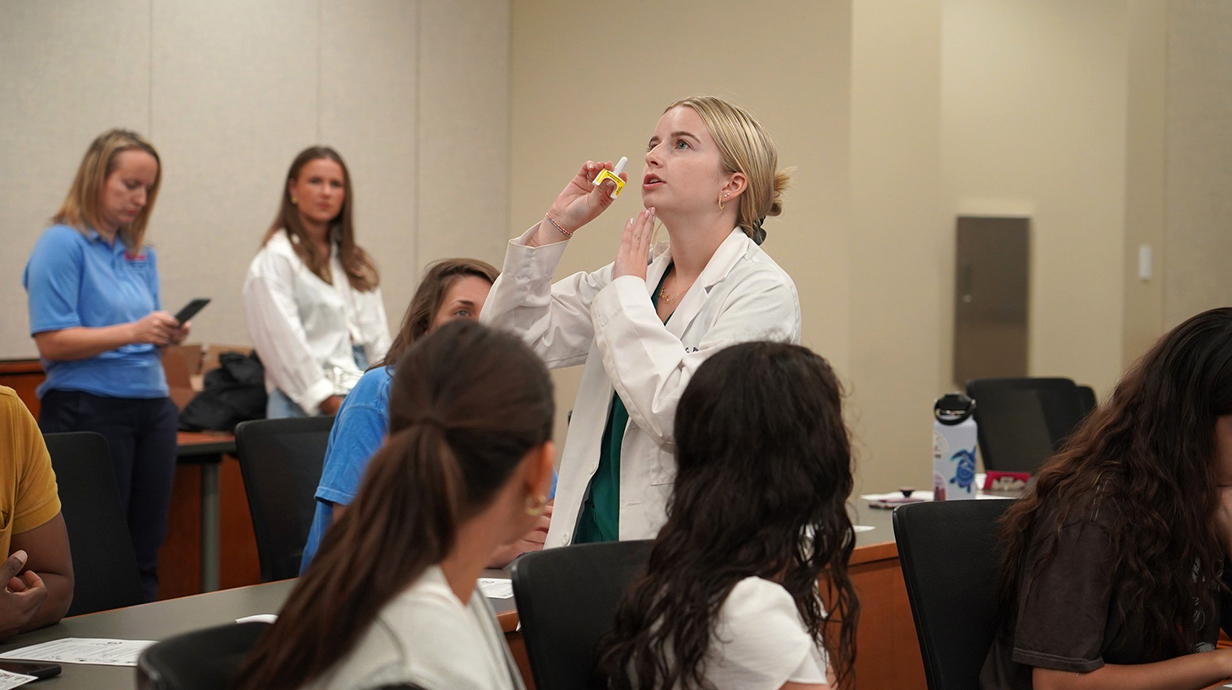 A young woman wearing a white lab coat demonstrates how to administer Narcan nasal spray for a room of seated teachers.