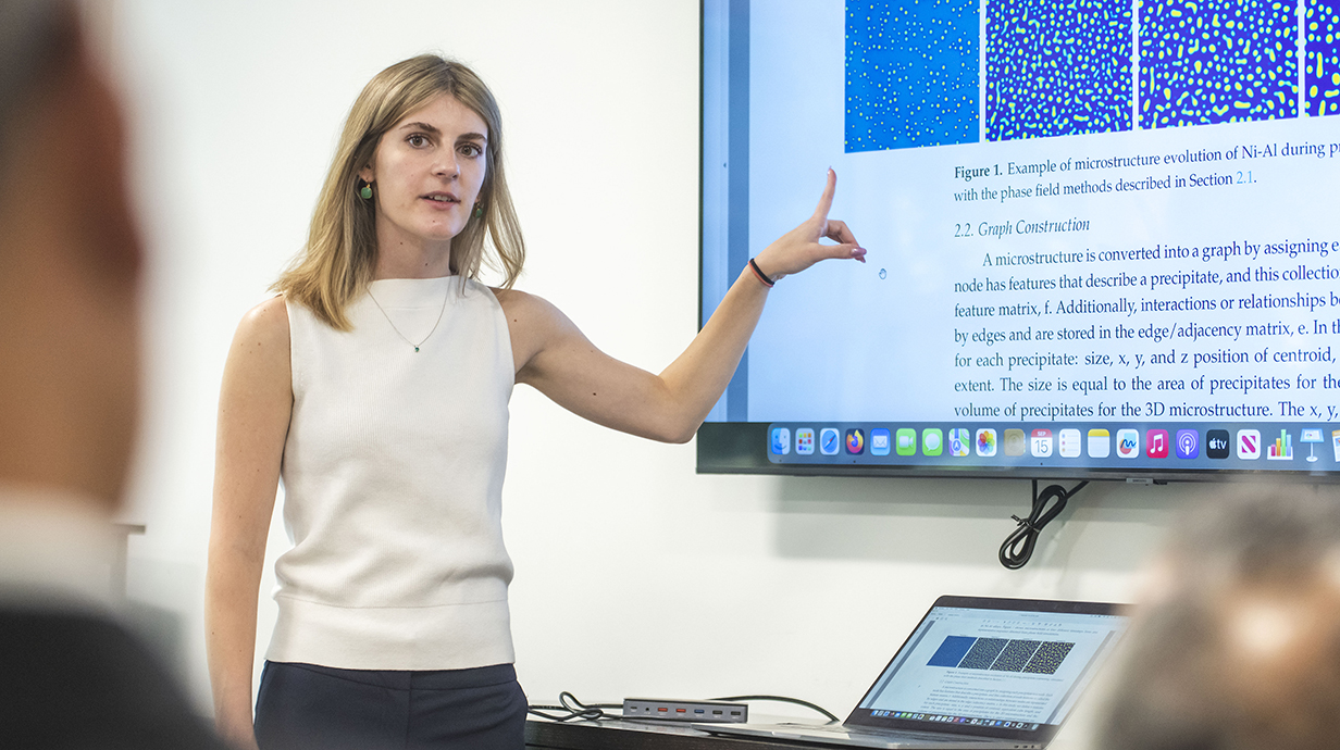 A young woman points to a diagram on a projection screen at the front of a classroom.