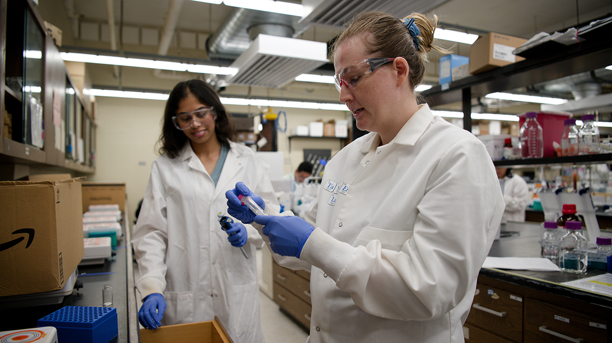 Two women wearing white lab coats and safety glasses work in a laboratory.