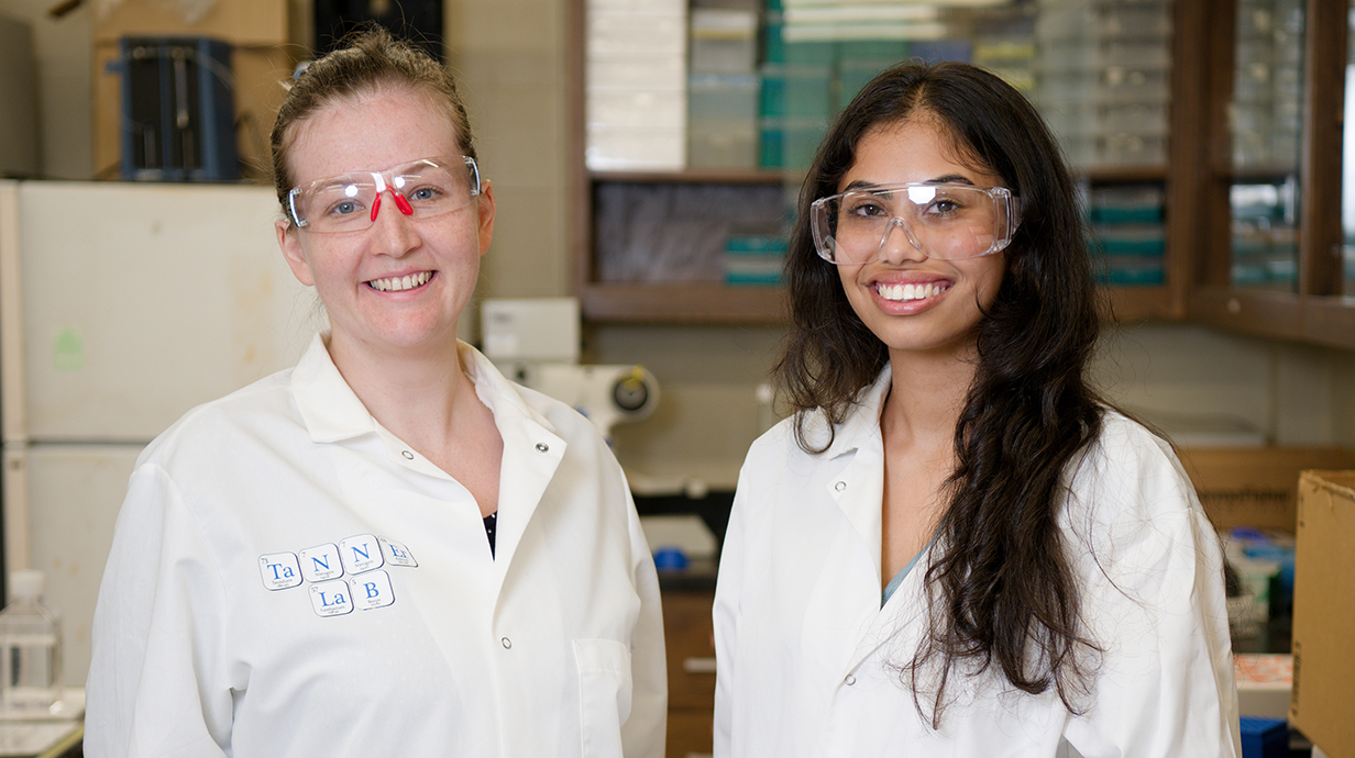 Two women wearing white lab coats and safety glasses stand in a laboratory.