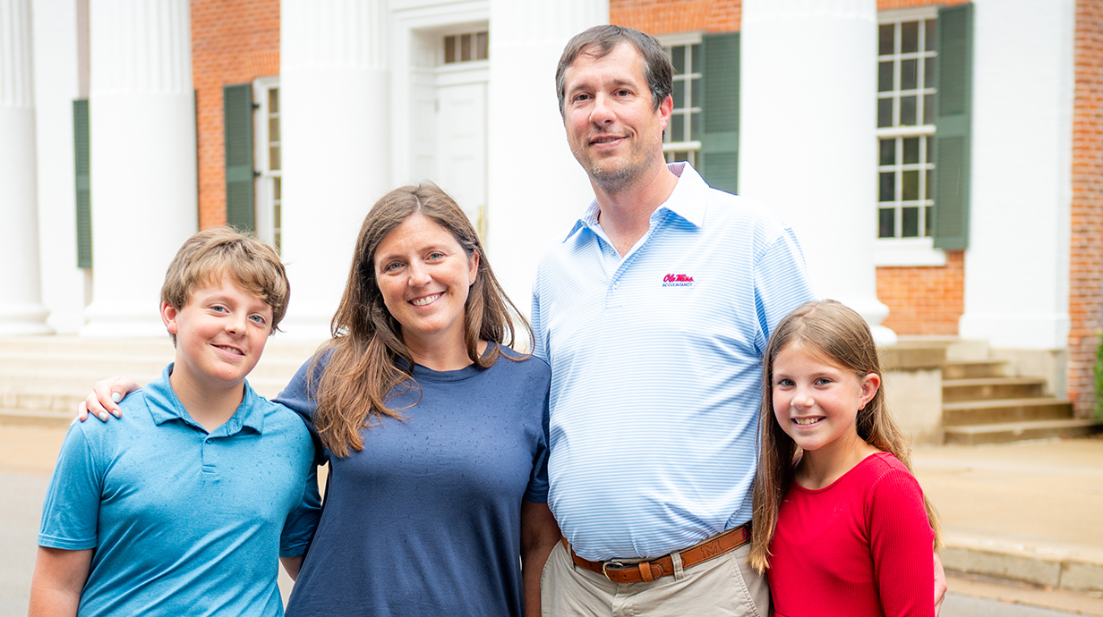 A man and woman pose for a family photo with a boy and girl in front of a white-columned building.