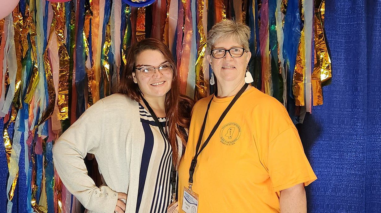 Two women pose for a photo in front of a backdrop covered in colorful streamers.