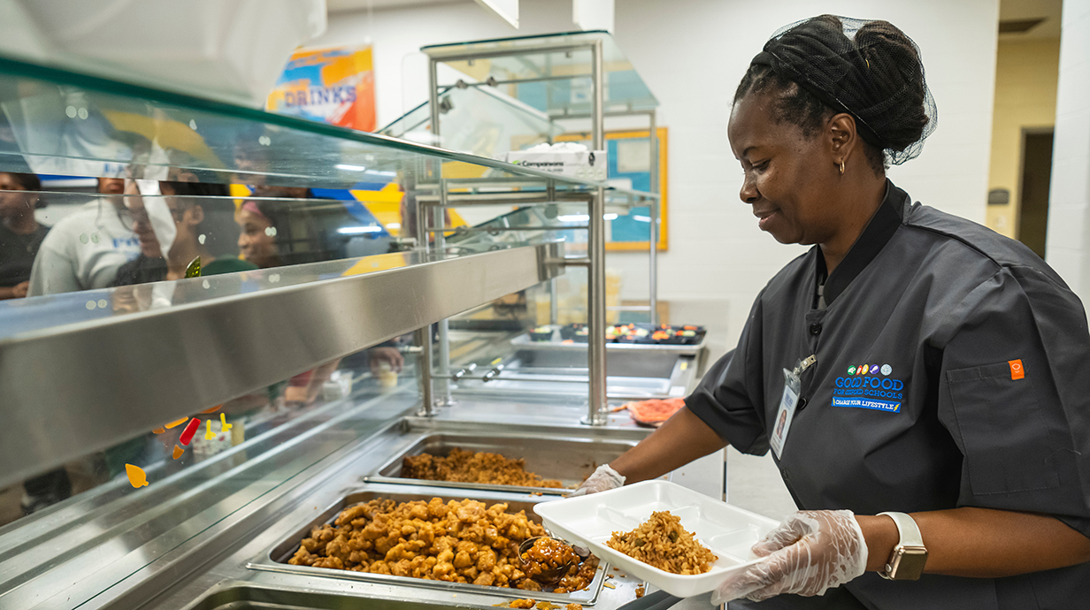 A woman working in a cafeteria line places food on a tray while customers wait on the other side of a glass barrier.