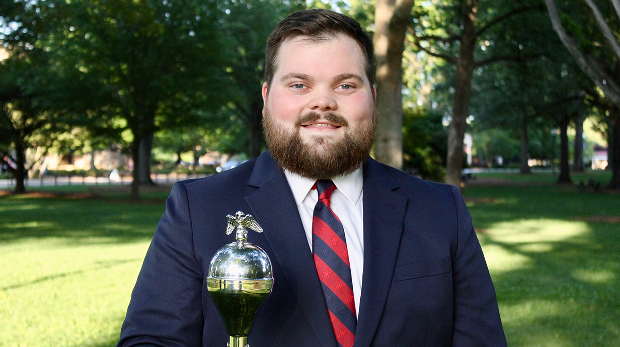 A young man wearing a suit holds a drum major's mace while standing in a park.