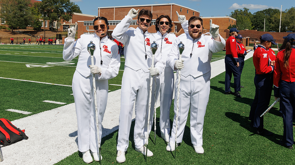 Four drum majors in white uniforms pose for a photo while marching band members in red and blue uniforms prepare to practice.