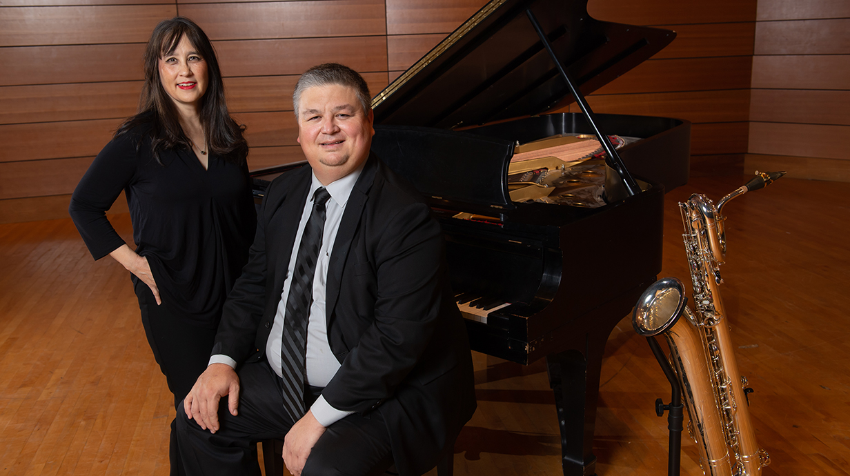 A woman stands beside a man sitting at a benches in front of a piano on a stage.