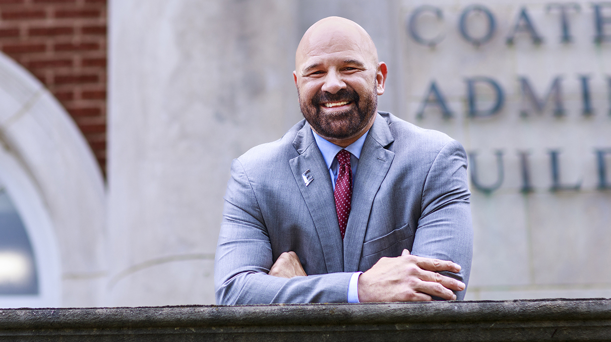 A man wearing a suit leans on a stone wall in front of a brick building.