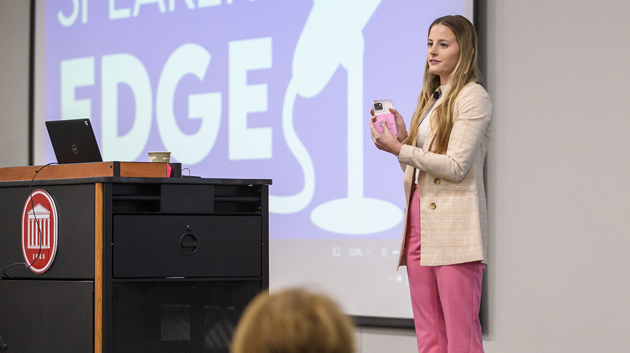 A young woman wearing business attire holds a cellphone while standing behind a lectern on a stage with a projection screen.