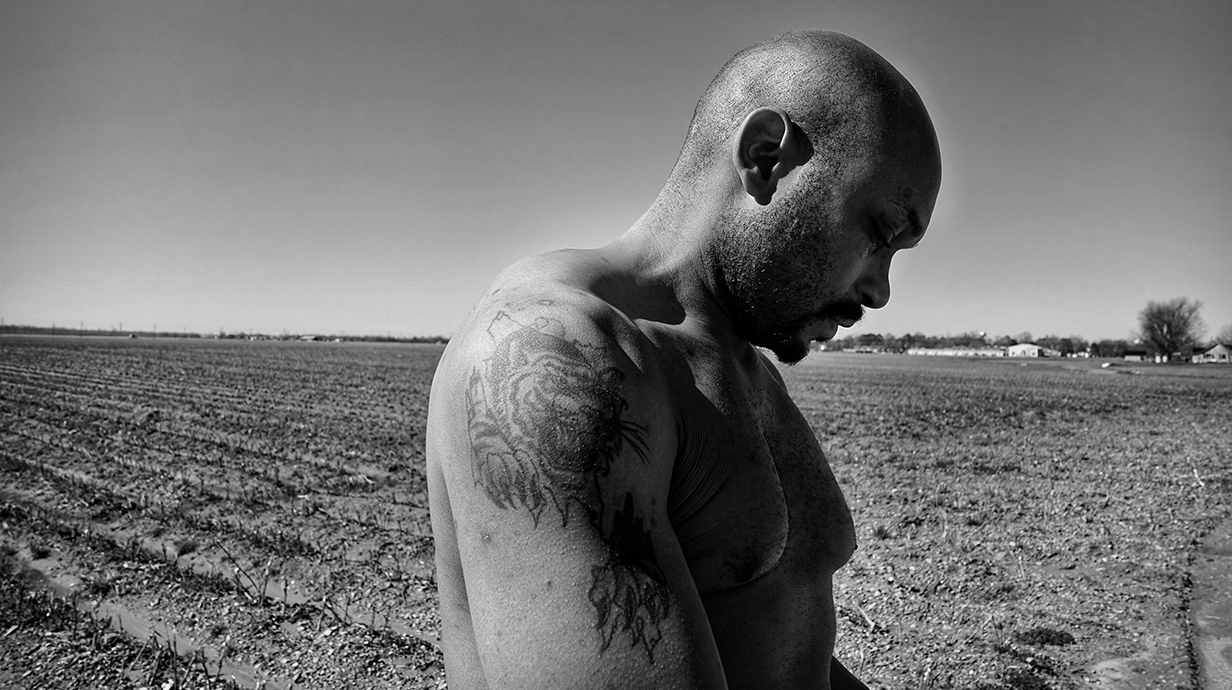 A shirtless man stands in a plowed field on a sunny day.