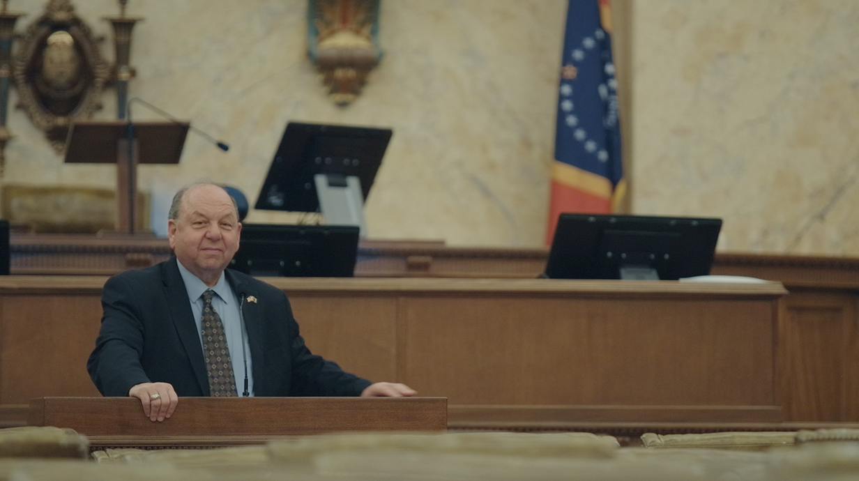 A man wearing a tan suit looks out over an empty legislative chamber.