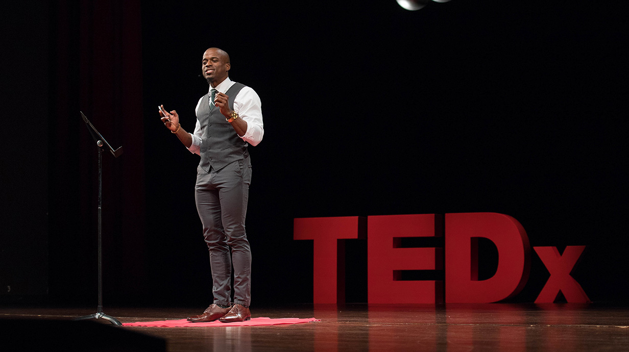 A man speaks on a darkened stage in front of giant red letters reading 'TEDx.'