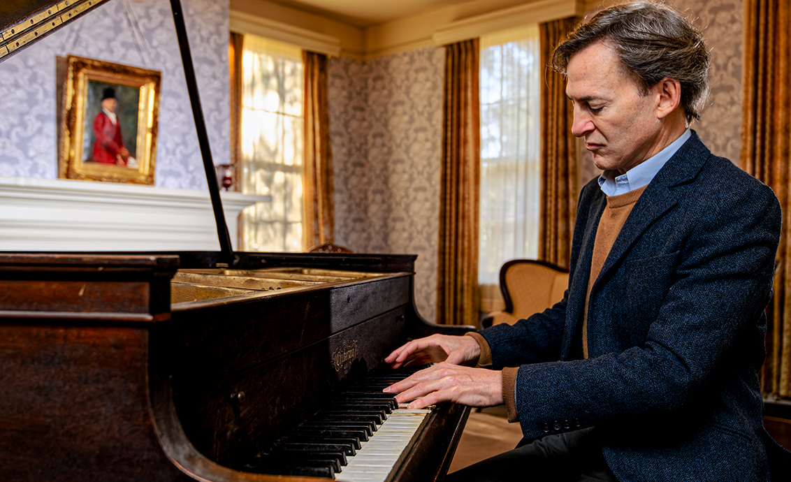 A man wearing a suit plays a piano in an ornate parlor.