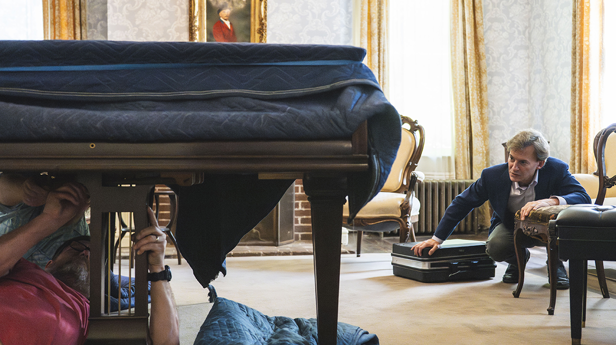 A man watches as two men work underneath a grand piano.