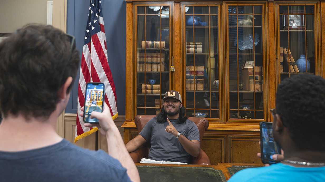 A young man sits at an ornate desk and flashes a thumbs-up sign as other young people take his photo.