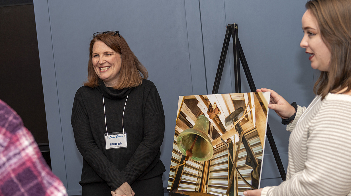 Two women show off a photo of an old bell in a hallway.