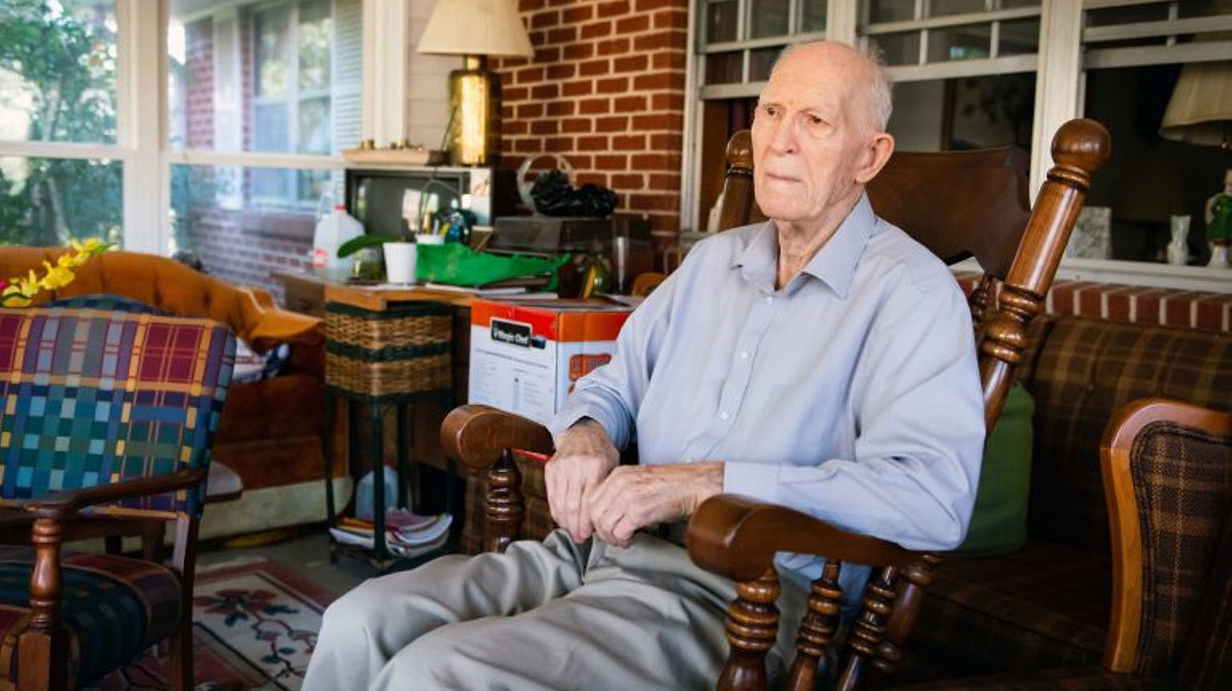 An elderly man sits in a rocker in a furnished sun porch.