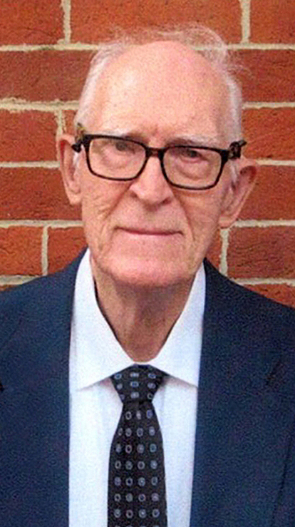 Headshot of an older man wearing a suit standing in front of a red brick wall.