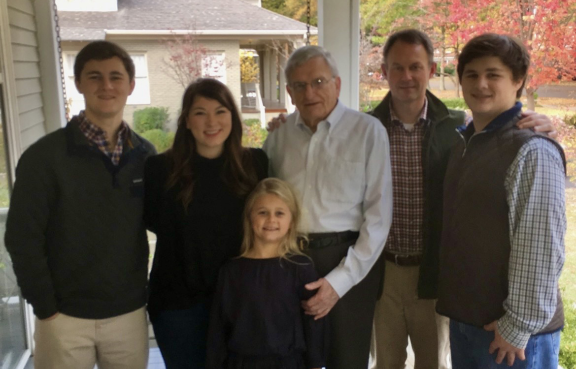 A group of people stands on a covered porch with fall colors behind them.