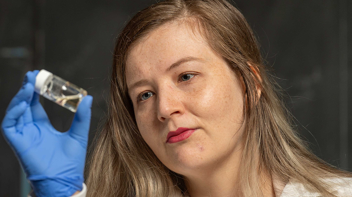 A woman wearing a white lab coat examines a small vial of liquid.