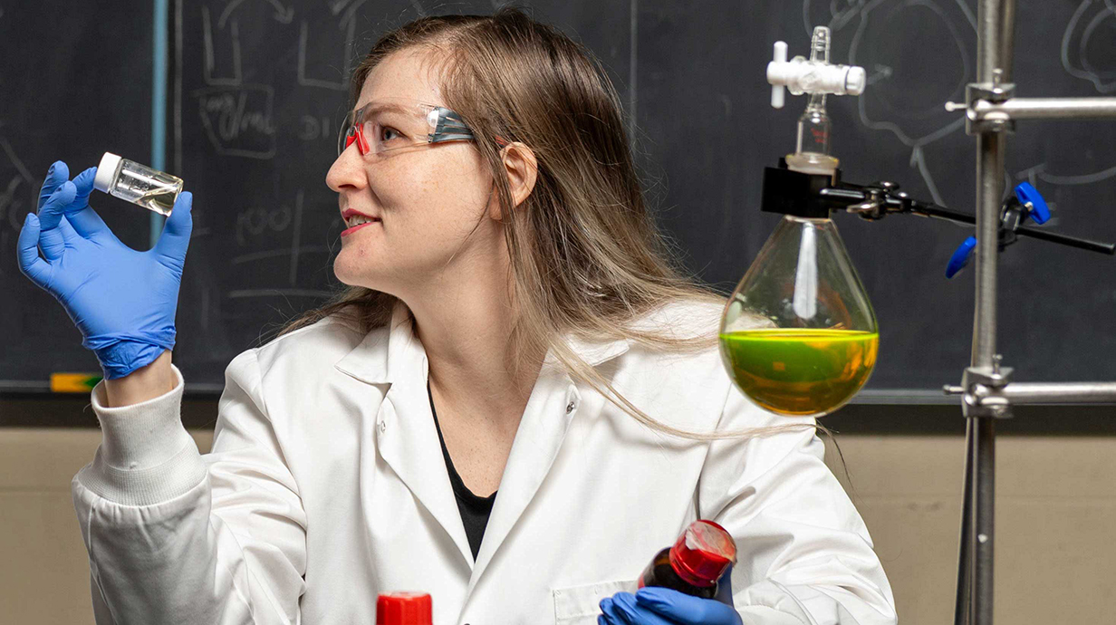A woman wearing a white lab coat and safety glasses holds a small vial of liquid in a laboratory.
