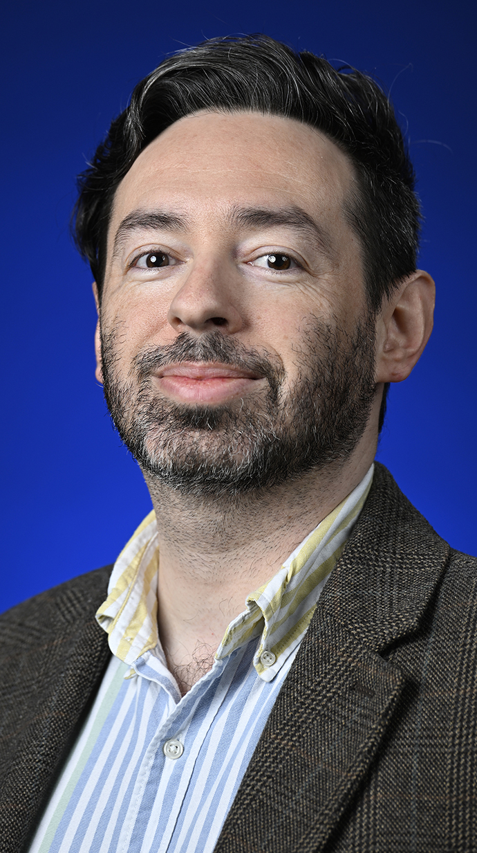 Headshot of a bearded man wearing a tweed jacket over a blue striped shirt.