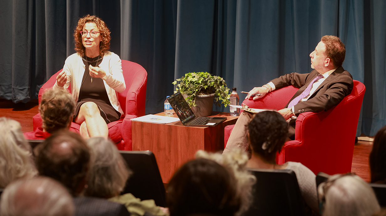A woman talks to an audience while sitting on a stage in an auditorium across from a man.