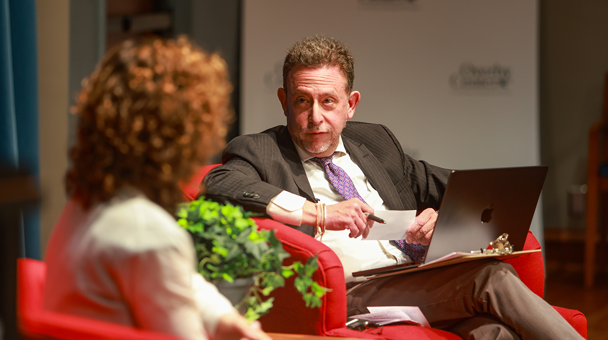 A man sitting on a stage speaks to a woman seated across from him.