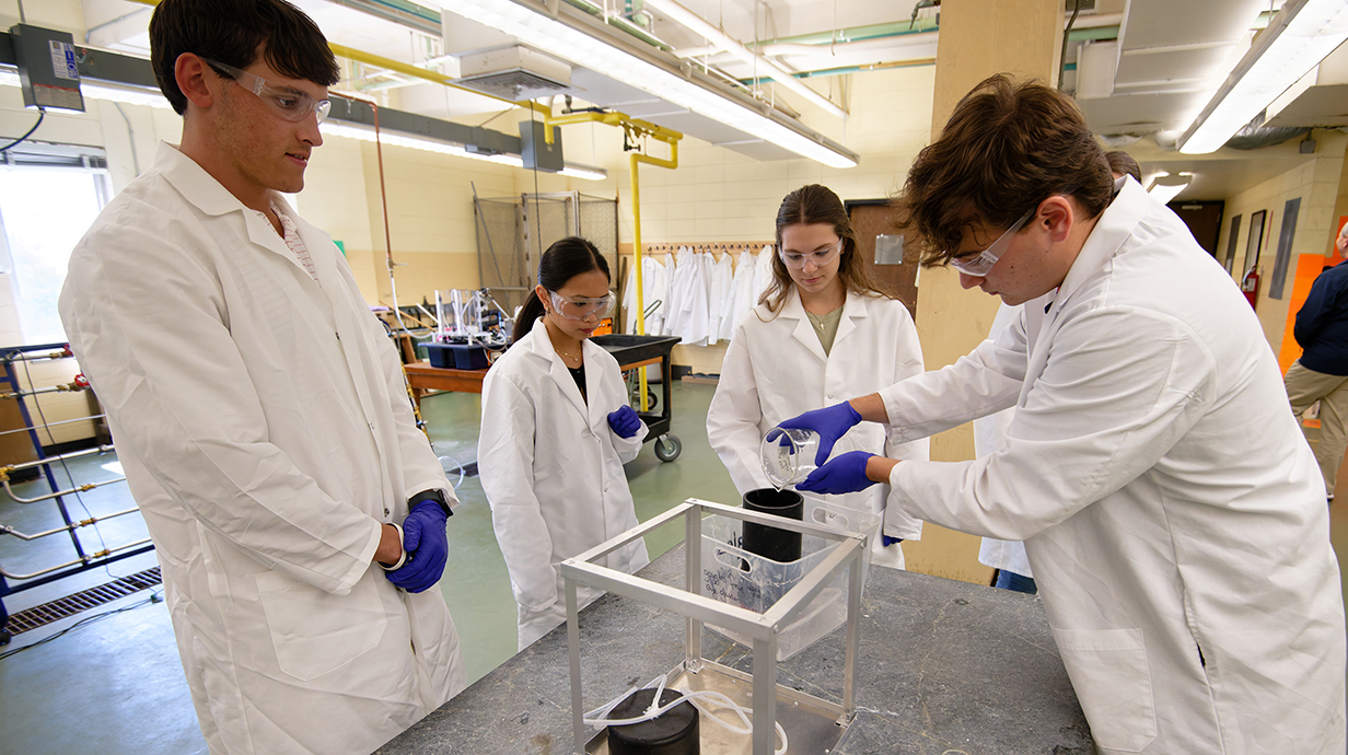 A young man wearing a white lab coat and safety glasses pours liquid from a beaker into a device while three other young people, all wearing lab coats and safety glasses, watch.