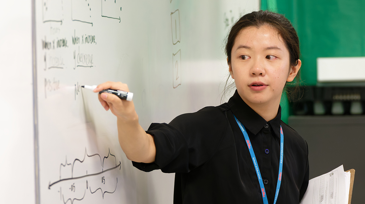 A young woman points to equations written on a whiteboard at the front of a classroom.