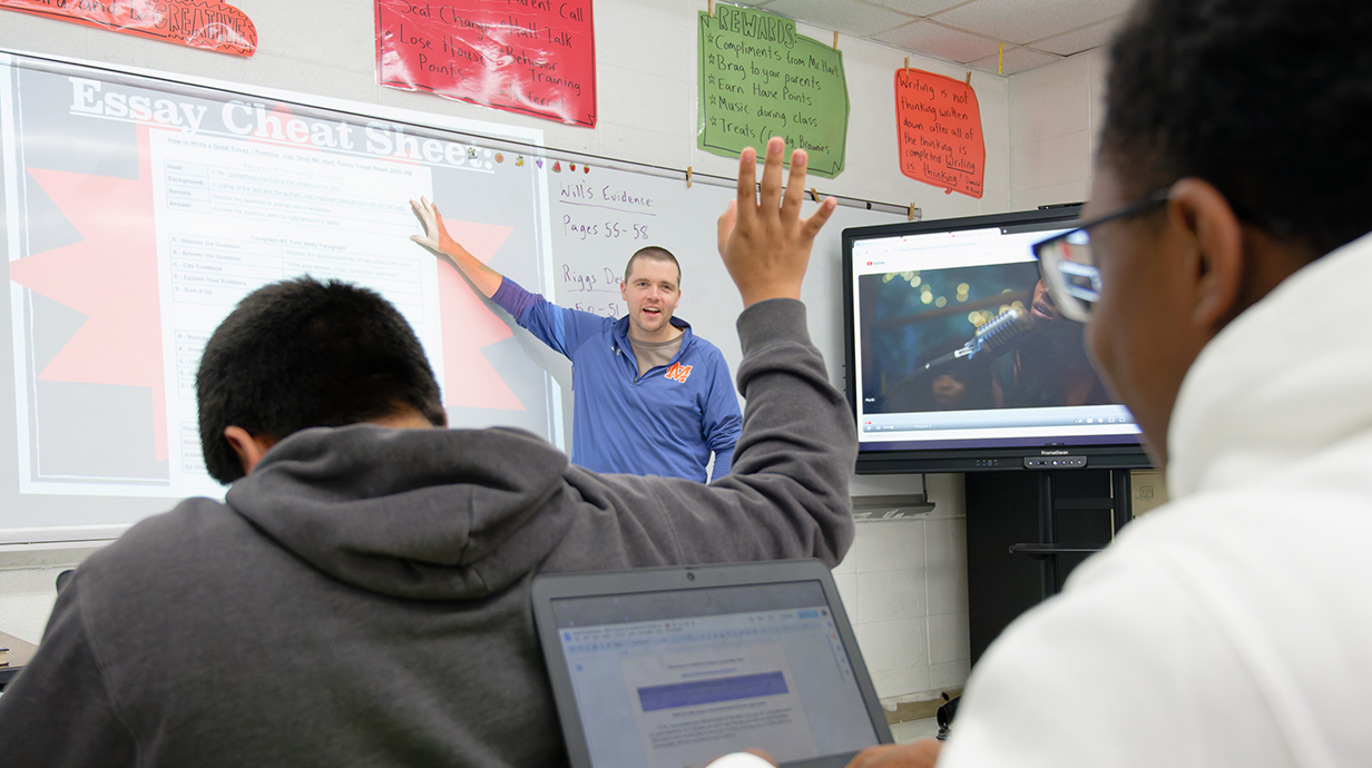 A tmale teacher points to a whiteboard in a classroom as multiple students raise their hands.