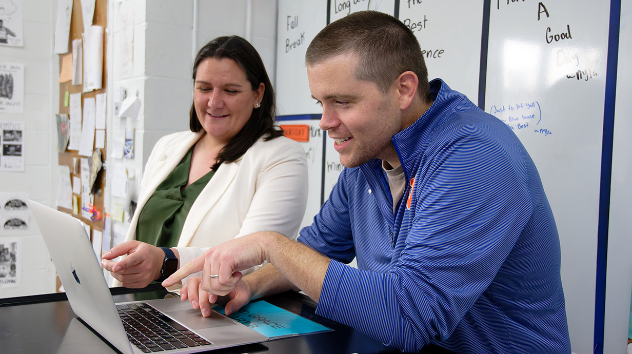 A woman and a man look at a laptop in a classroom.