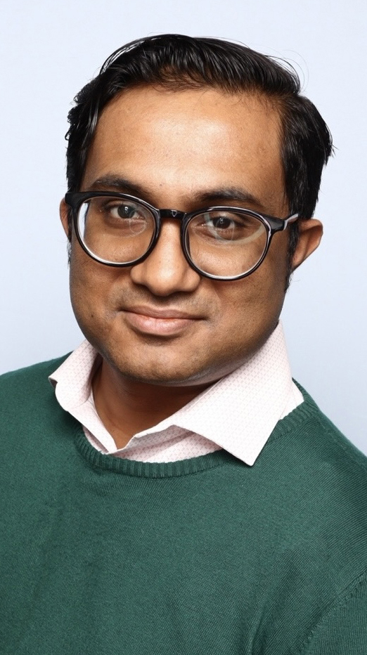 Headshot of a man wearing a green sweater over a white dress hirt.