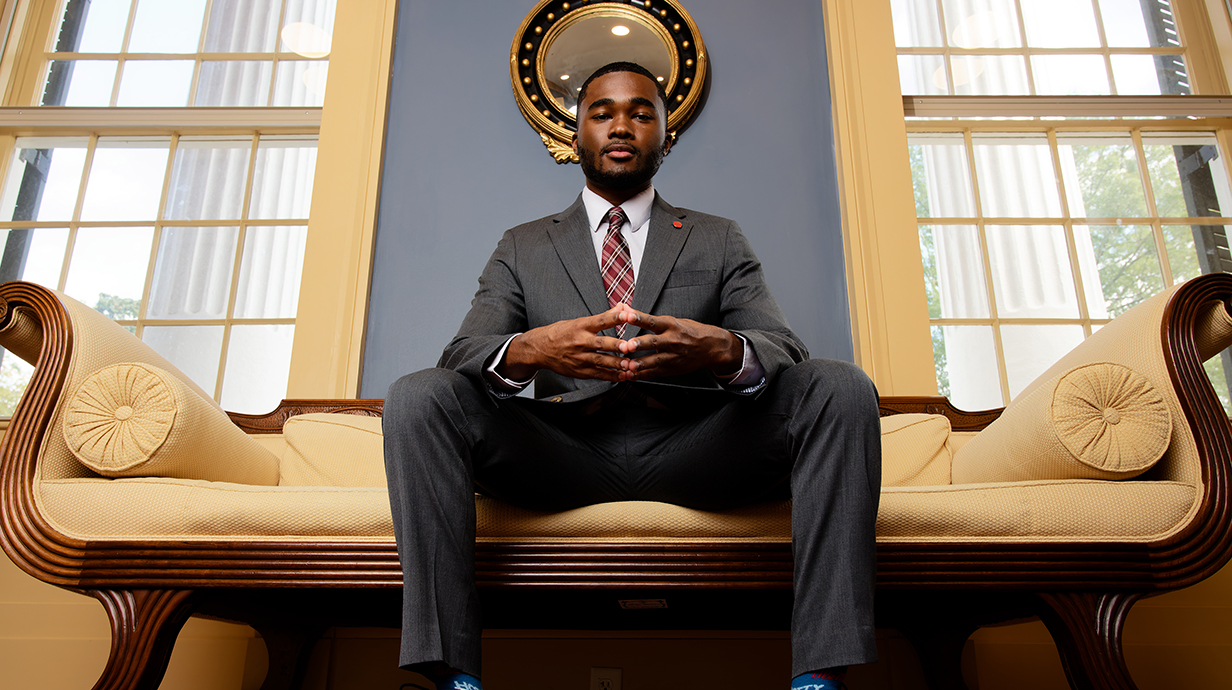 A young man wearing a suit sits on a couch in an ornate parlor looking down at the camera.