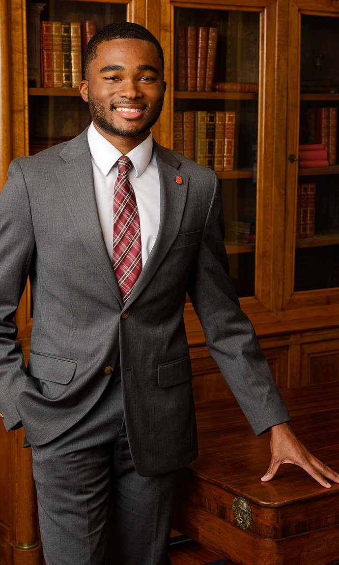 A young man wearing a suit stands beside an ornate bookcase.