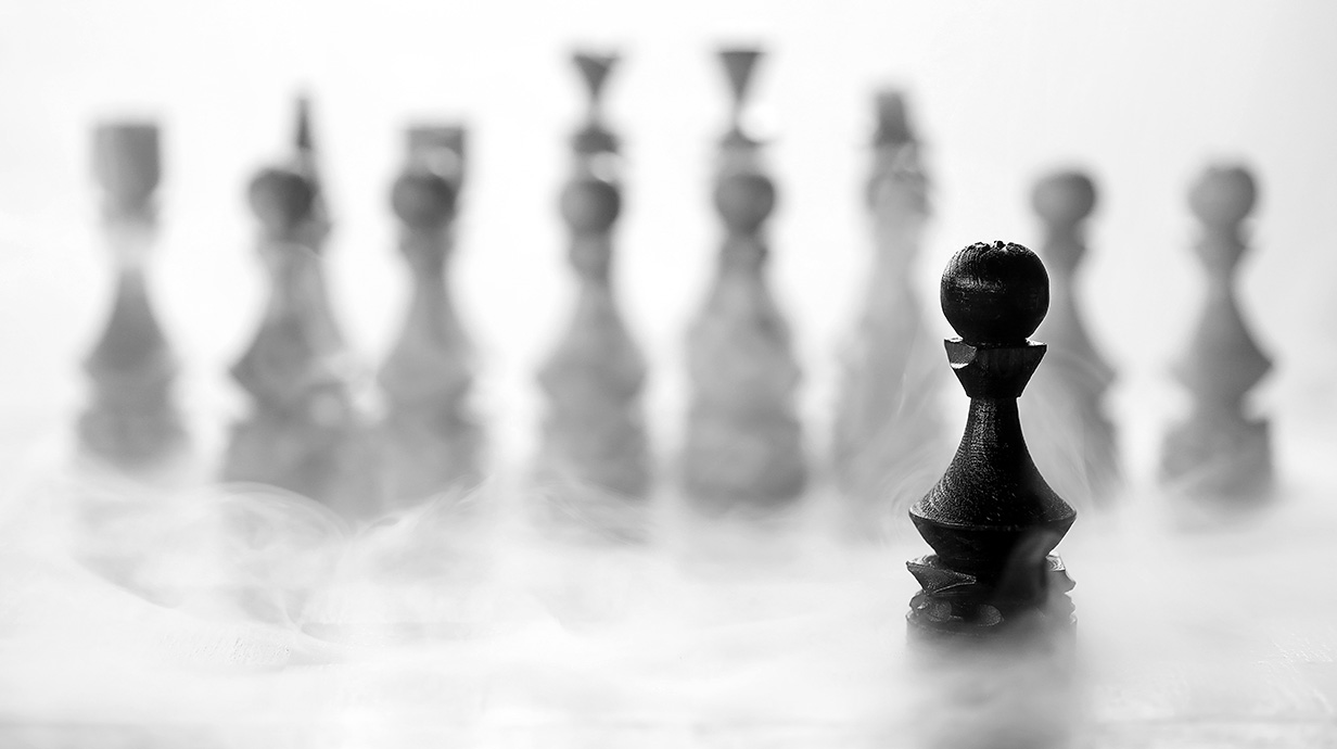 Photograph of a row of white chess pieces on a chess board, with a black pawn in the foreground.