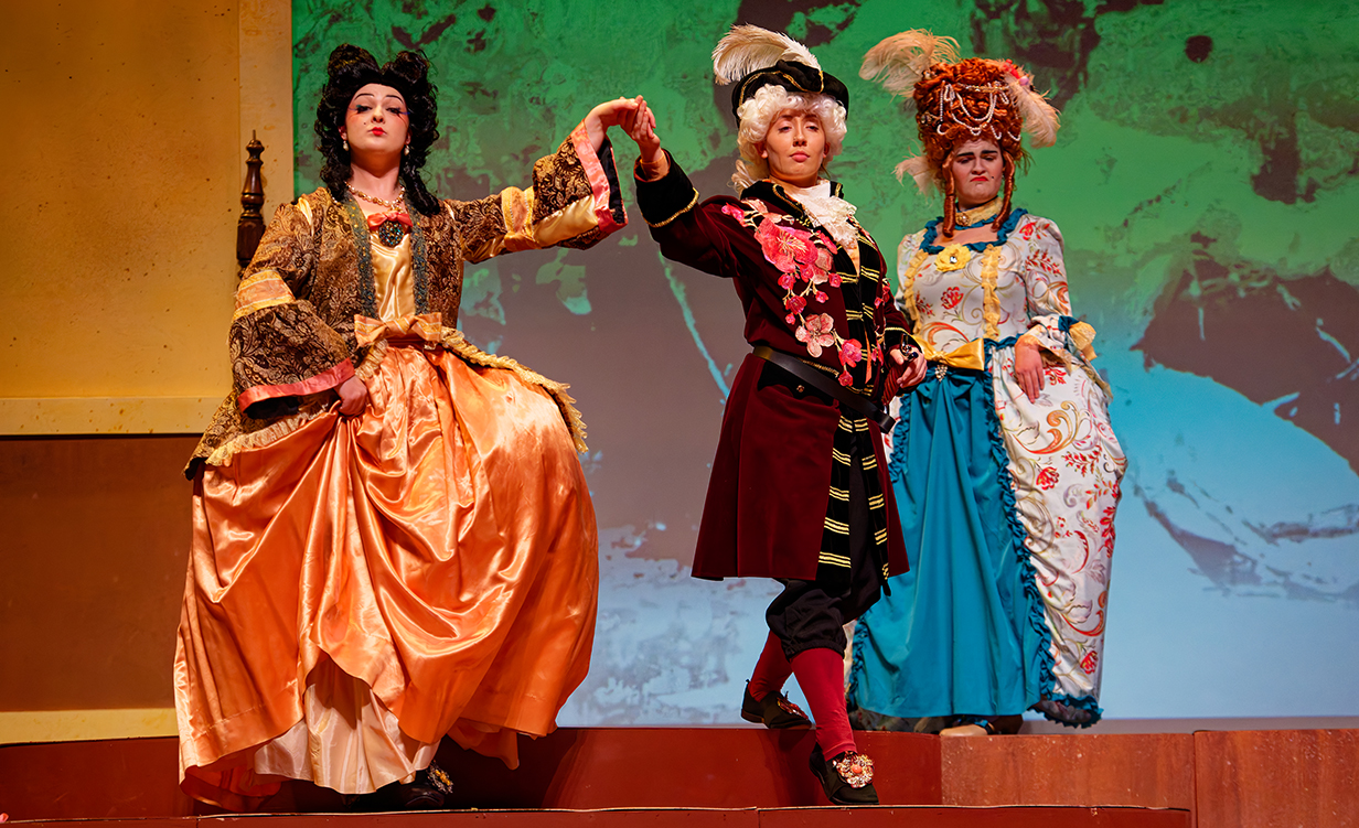 Three women wearing colorful costumes and theatrical makeup sing and dance on a stage.