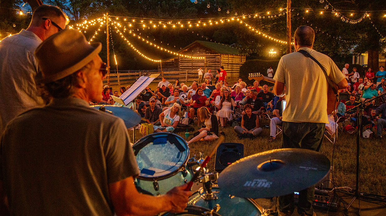 A band performs an outdoor concert at night before a crowd seated on a lawn.