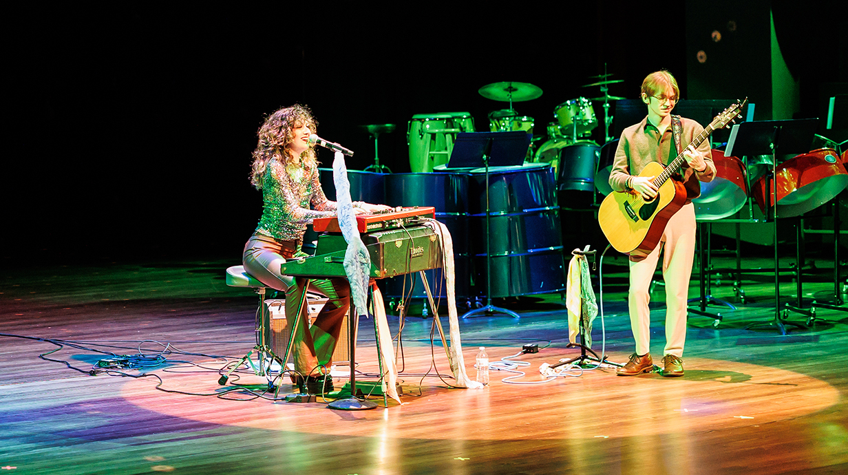 A woman plays a keyboard and sings and a man plays guitar on a stage.