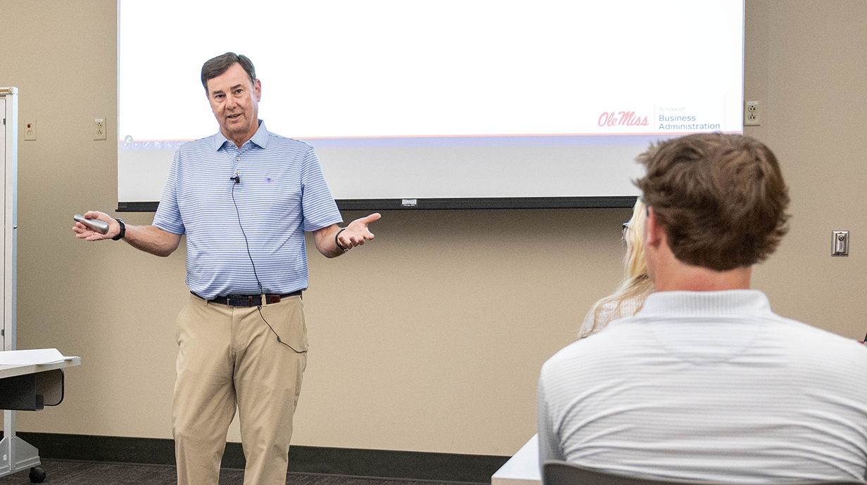A man standing in front of a projection screen in a classroom gestures to students seated nearby.