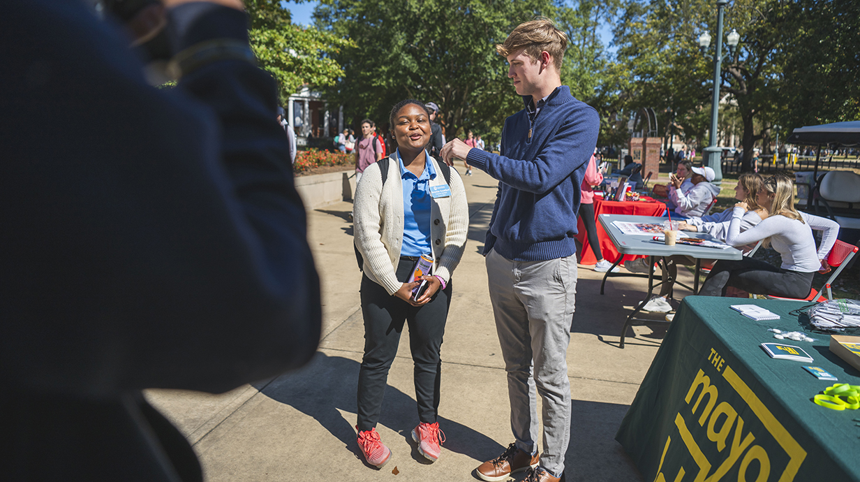 A young man with a microphone interviews a young woman on an outdoor plaza lined with tables.