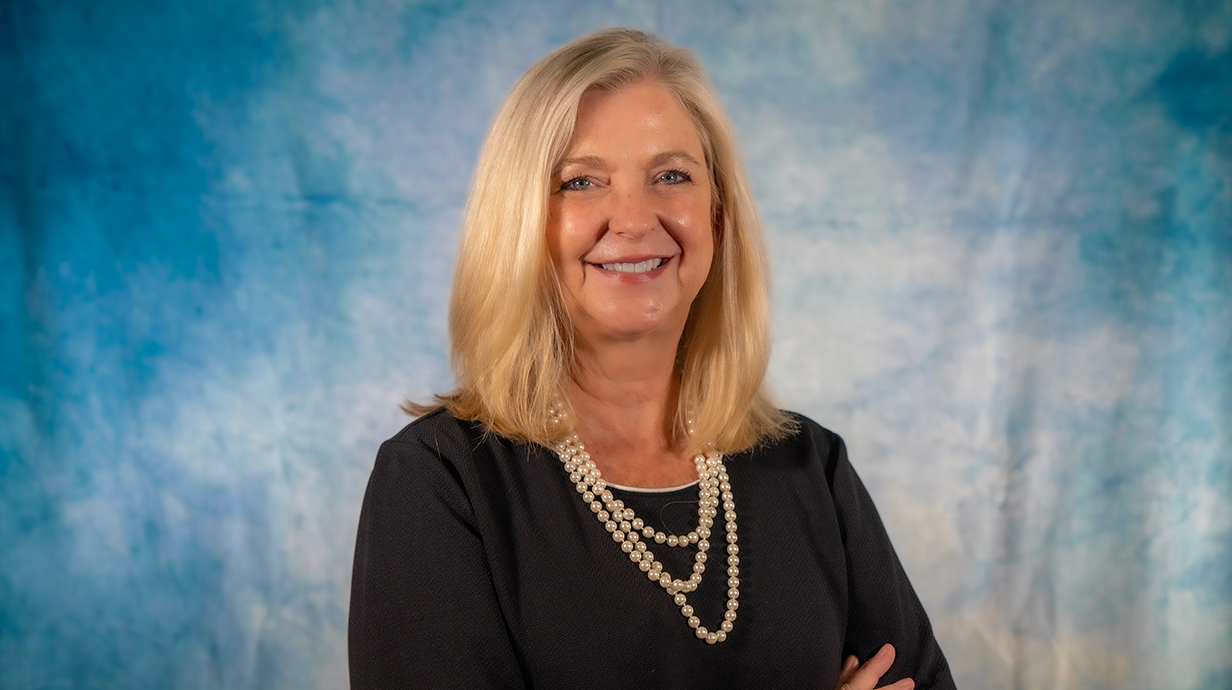 A woman wearing a dark dress and pearls stands in front of a light blue backdrop.