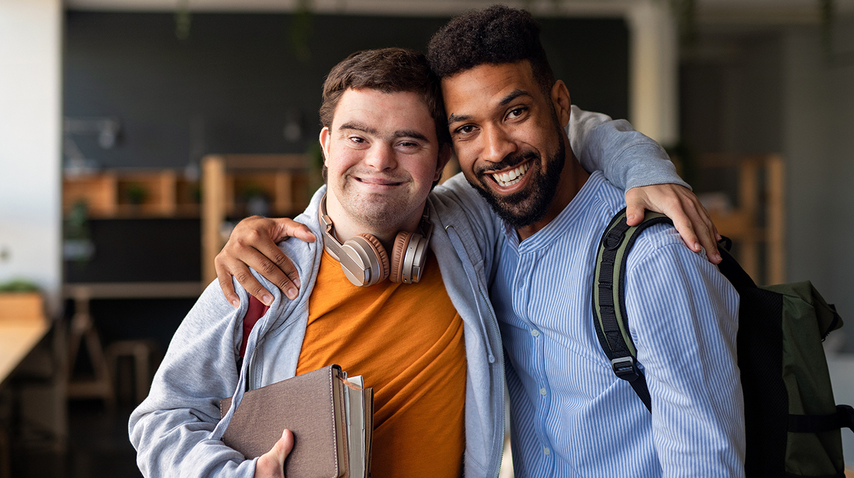 A man stands with his arm around another man with Down syndrome.