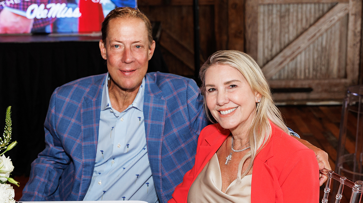 A man and a woman sit together at a table with a projection screen behind them.