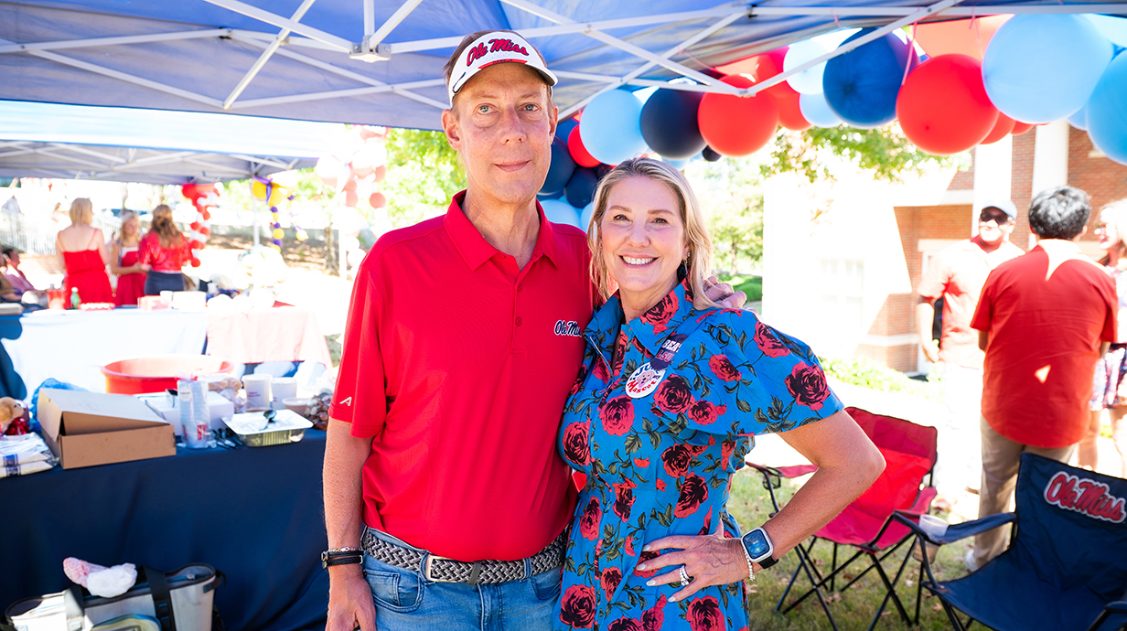 A man and a woman stand together underneath a pop-up tent lined with red-and-blue balloons and tables of food.