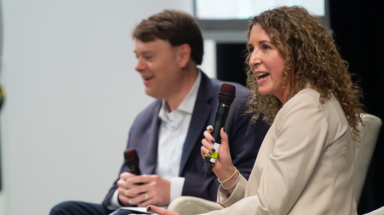 A woman speaks into a microphone while sitting on a stage next to a man.