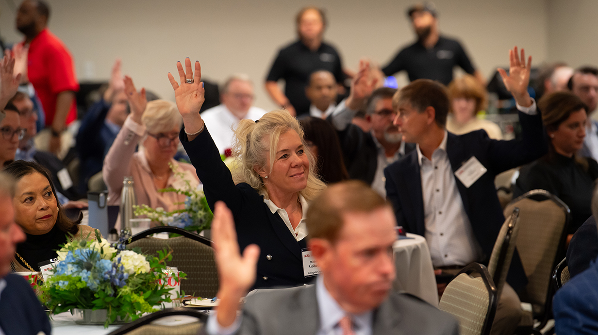 Several people raise their hands while seated in a crowded conference hall.