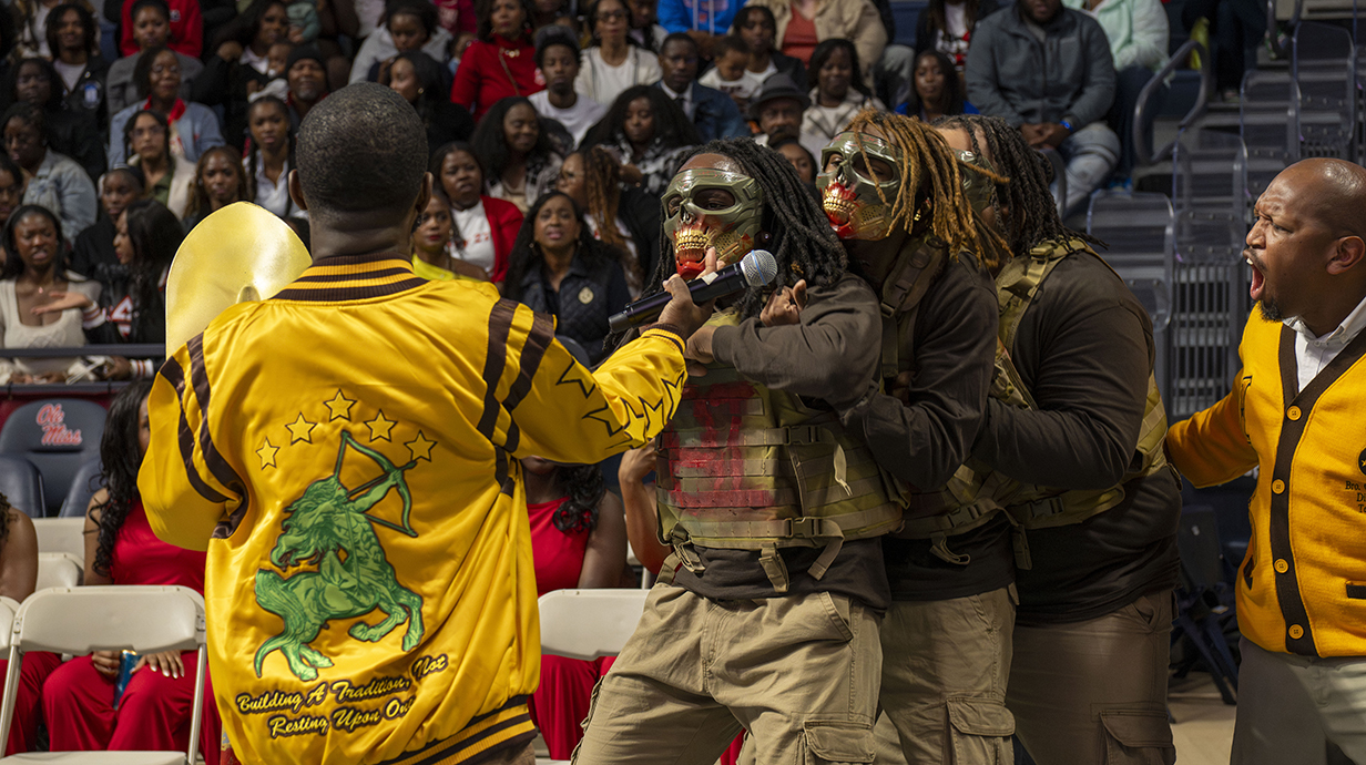 Several young men wearing masks conduct a ceremony in an arena as other young men urge them on.