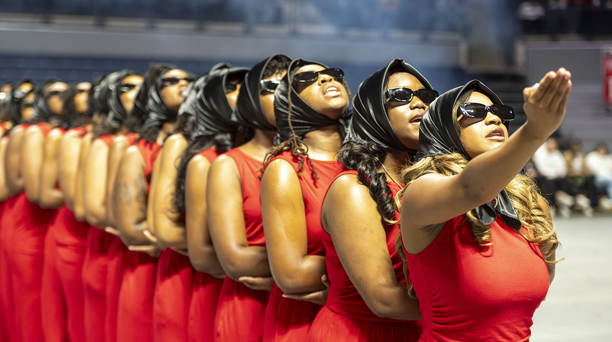 A group of young women, all wearing red dresses and black sunglasses, perform a dance in an arena.